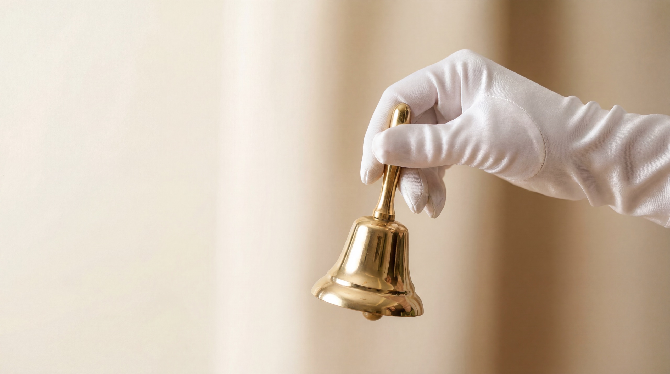 A white-gloved hand ringing a polished gold bell against a cream backdrop